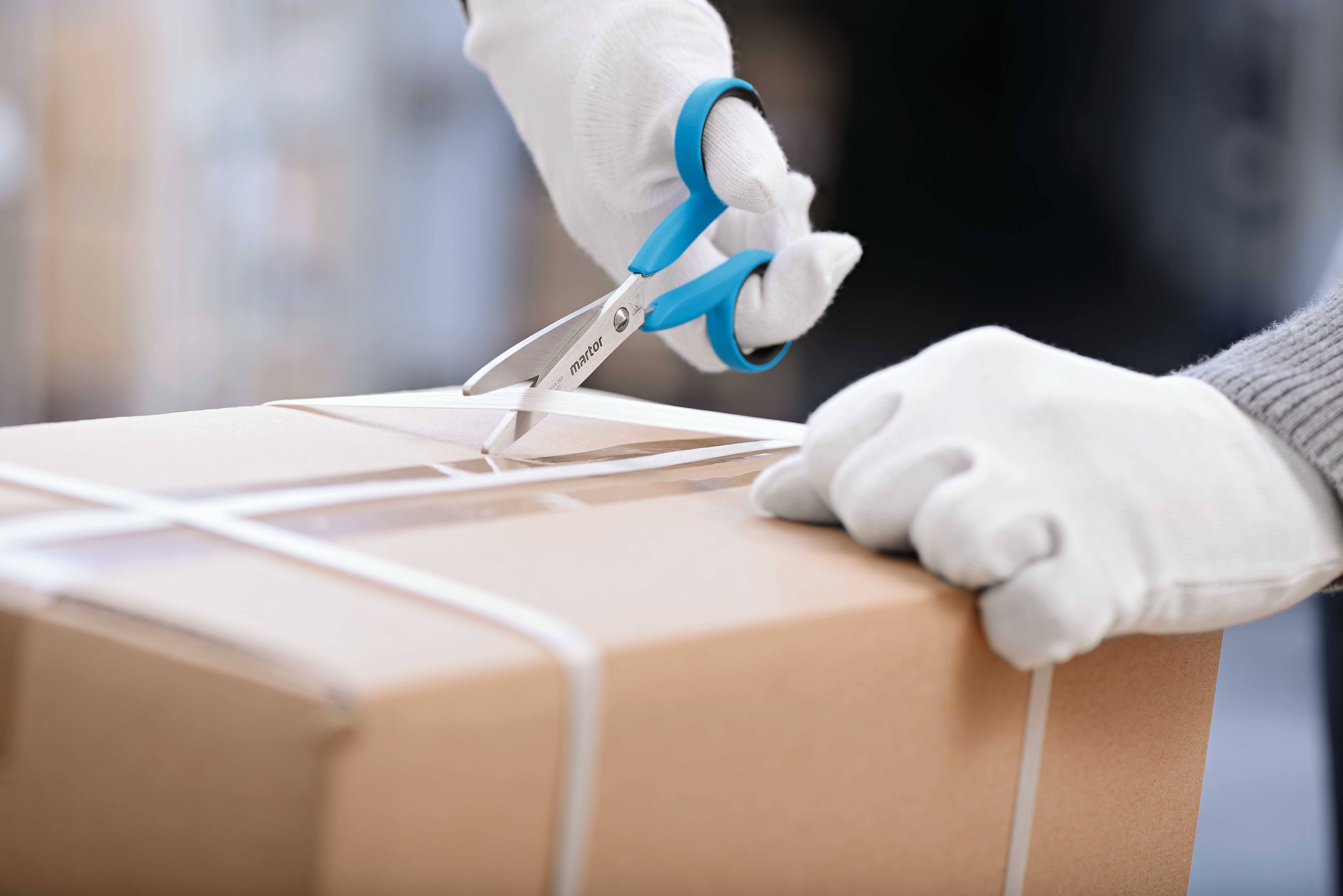 Une personne coupe un ruban d'emballage d'un carton à l'aide de ciseaux. Les mains portent des gants blancs.