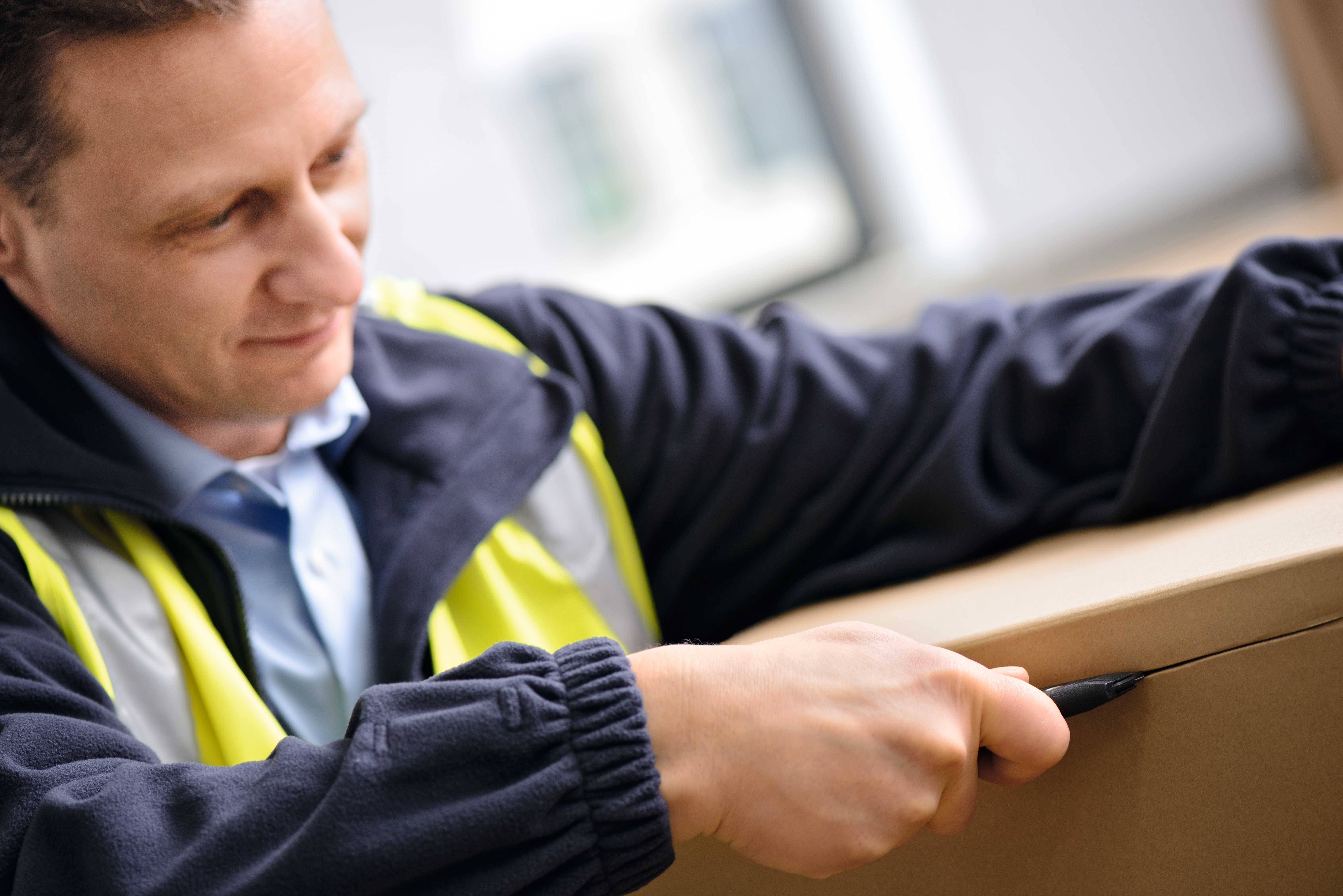 Un homme en tenue de travail ouvre prudemment un carton à l'aide d'un cutter.