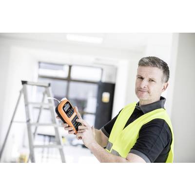 Un homme portant un gilet de sécurité jaune utilise un appareil portable dans une pièce lumineuse avec une échelle en arrière-plan. Il regarde l'appareil photo.