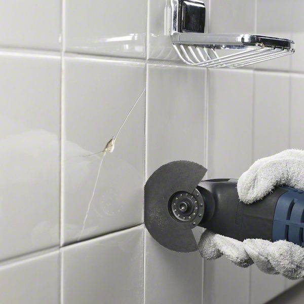 Une personne utilise un outil électrique avec un petit disque à tronçonner pour retirer des joints endommagés dans une salle de bain aux carrelages blancs.