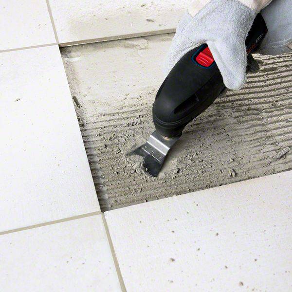 Un outil retire les anciennes joints de carrelage d'un sol. Une main tient l'appareil, tandis que des restes de mortier détachés sont visibles.