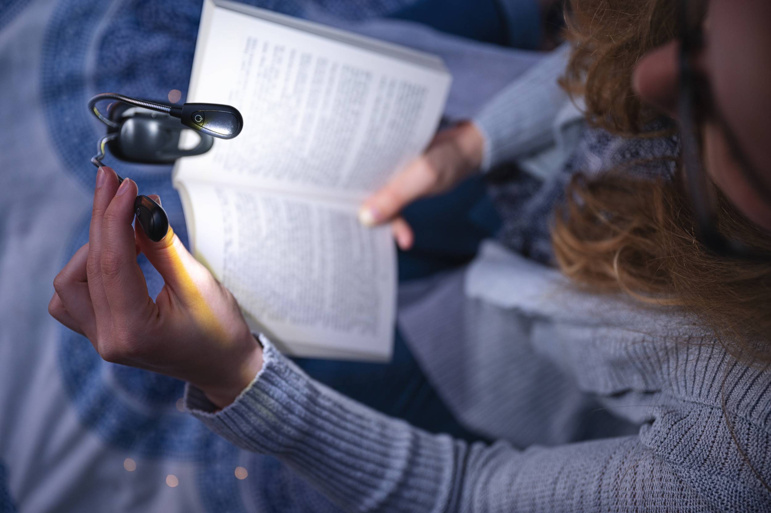 Une personne lit un livre dans l'obscurité avec une petite lampe de lecture fixée sur le livre. La scène semble calme et concentrée.