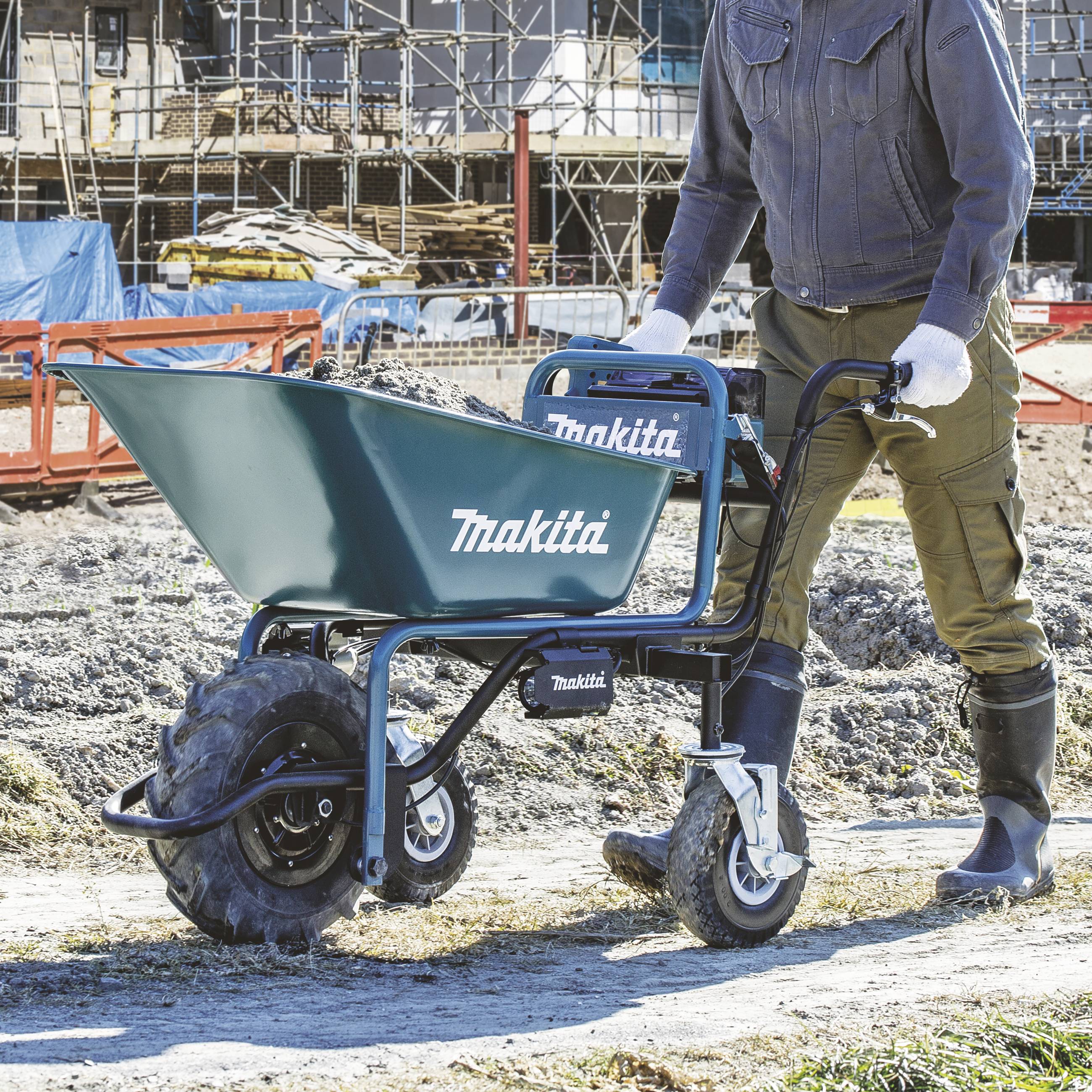 Une personne pousse une brouette bleue Makita à deux roues sur un chantier de construction. À l'arrière-plan, on aperçoit des matériaux de construction.