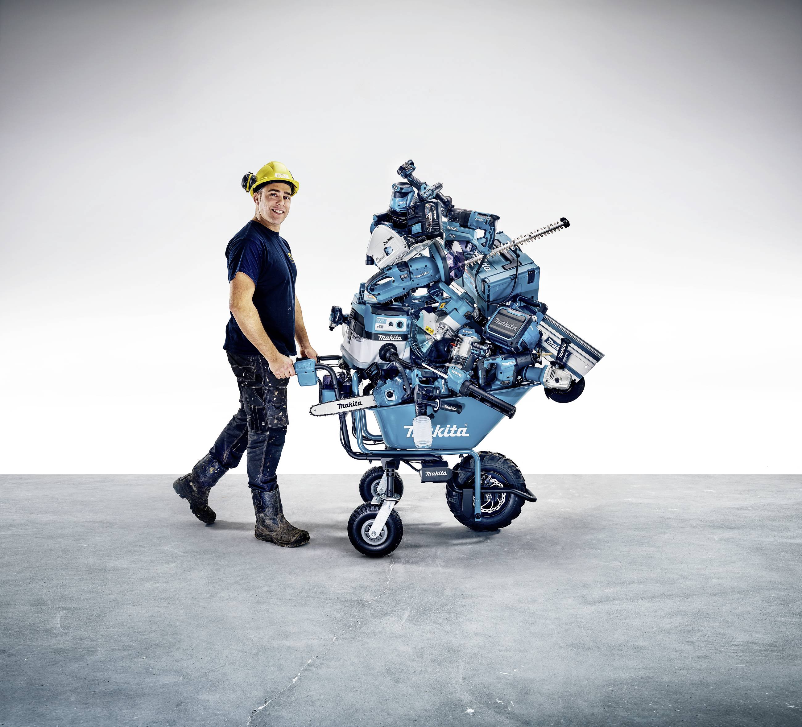 Un ouvrier souriant pousse une brouette remplie d'outils et de machines sur un sol en béton.