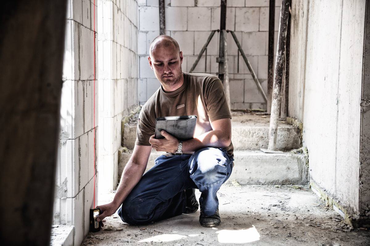 Un homme est agenouillé dans un bâtiment en construction et mesure les murs avec un appareil. Il tient une planche à pince et porte des vêtements de travail.