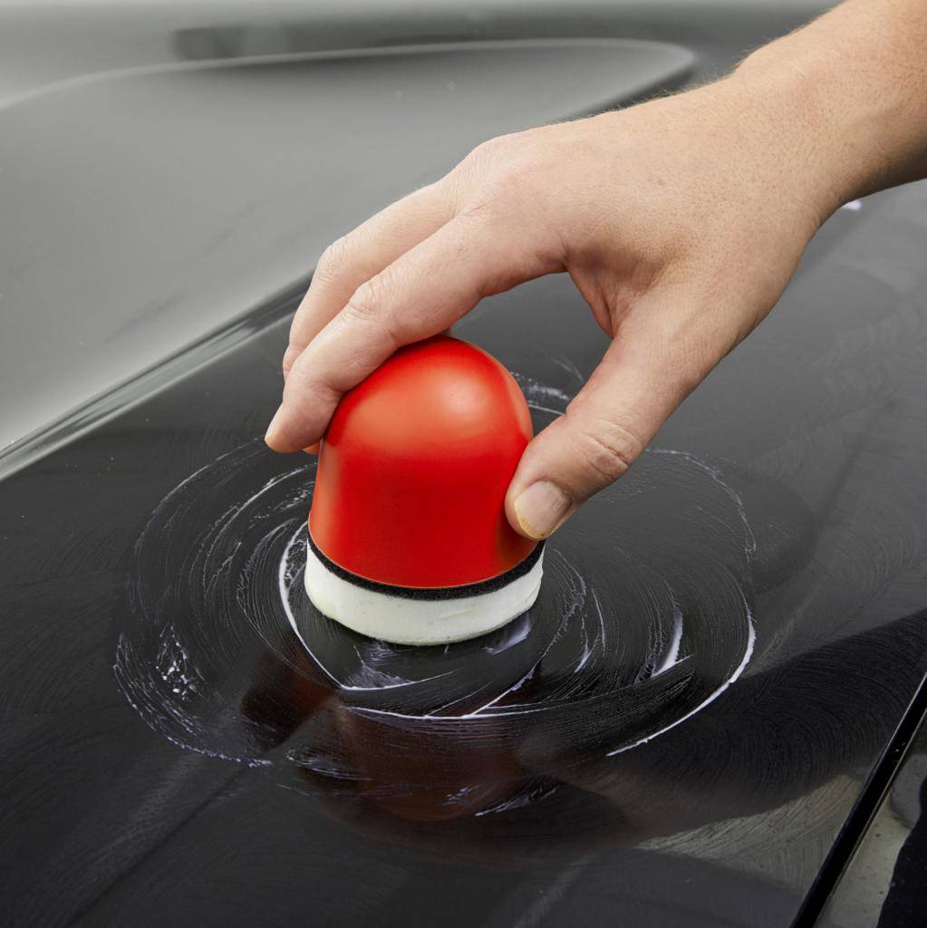 Une main tient un pad de polissage rouge qui est utilisé sur un vernis noir de voiture pour polir la surface.