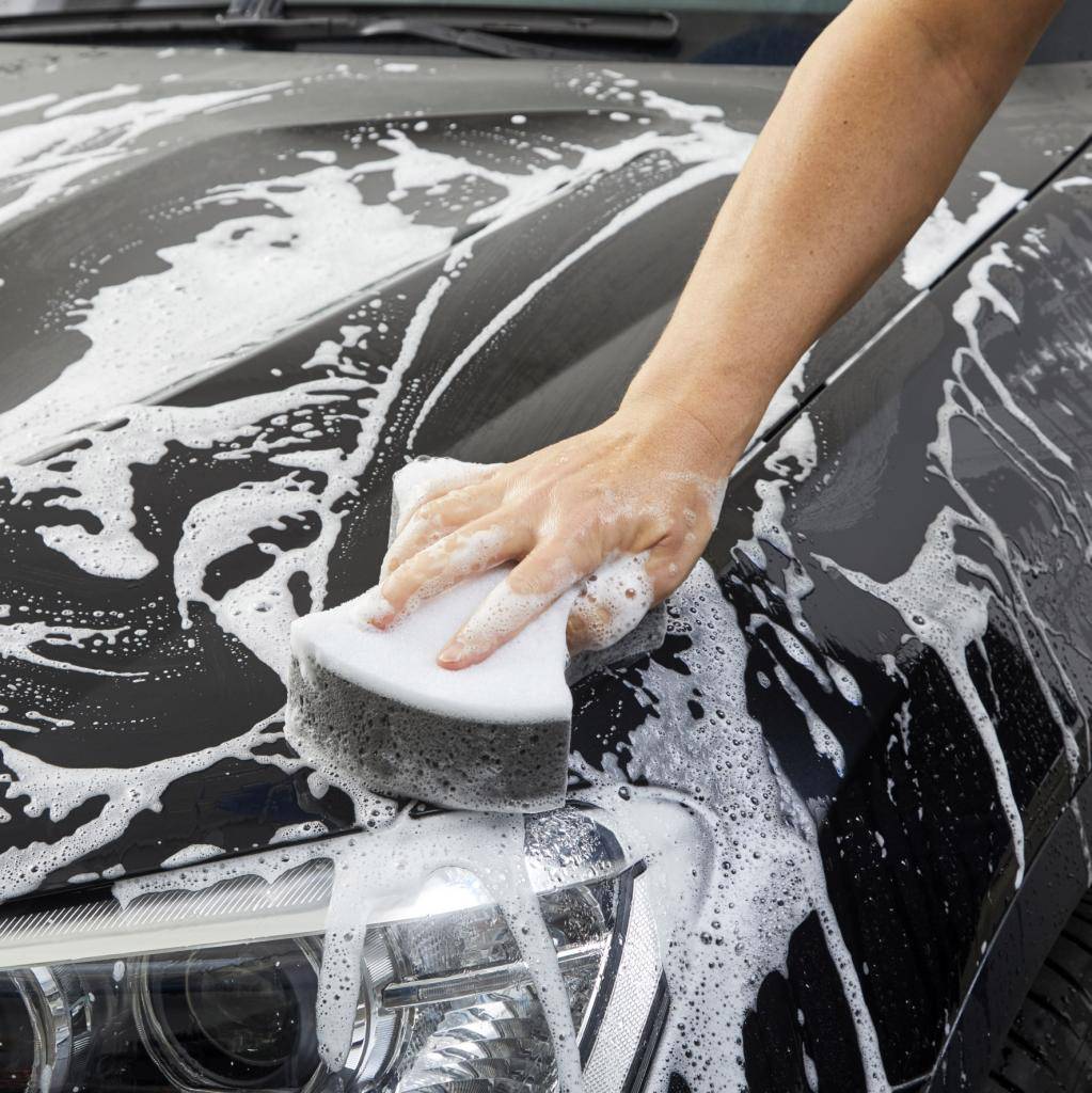 Une main nettoie une voiture avec une éponge, recouverte de mousse. L'accent est mis sur le lavage du capot et des phares.