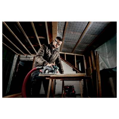 Une personne portant des lunettes de sécurité utilise une scie circulaire sur une planche de bois dans une pièce en construction partielle, avec des poutres apparentes et des outils visibles.