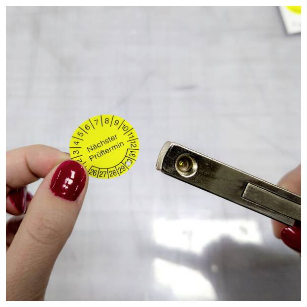 Une personne tient un autocollant de contrôle jaune portant l'étiquette « Nächster Prüftermin » avec des numéros et marqué par un poinçon, montrant une attention concentrée sur le contrôle technique du véhicule.