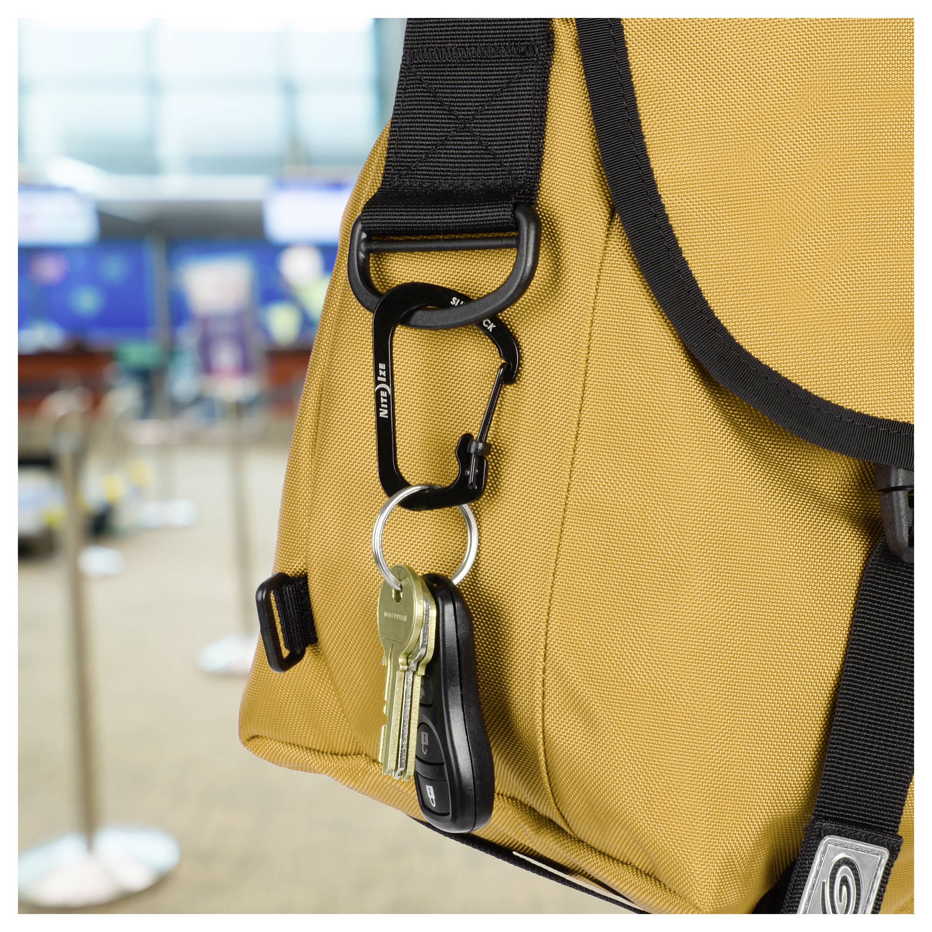 Un gros plan sur un sac jaune avec un mousqueton tenant des clés, dont une clé de voiture. Un comptoir d'enregistrement d'aéroport flou en arrière-plan.