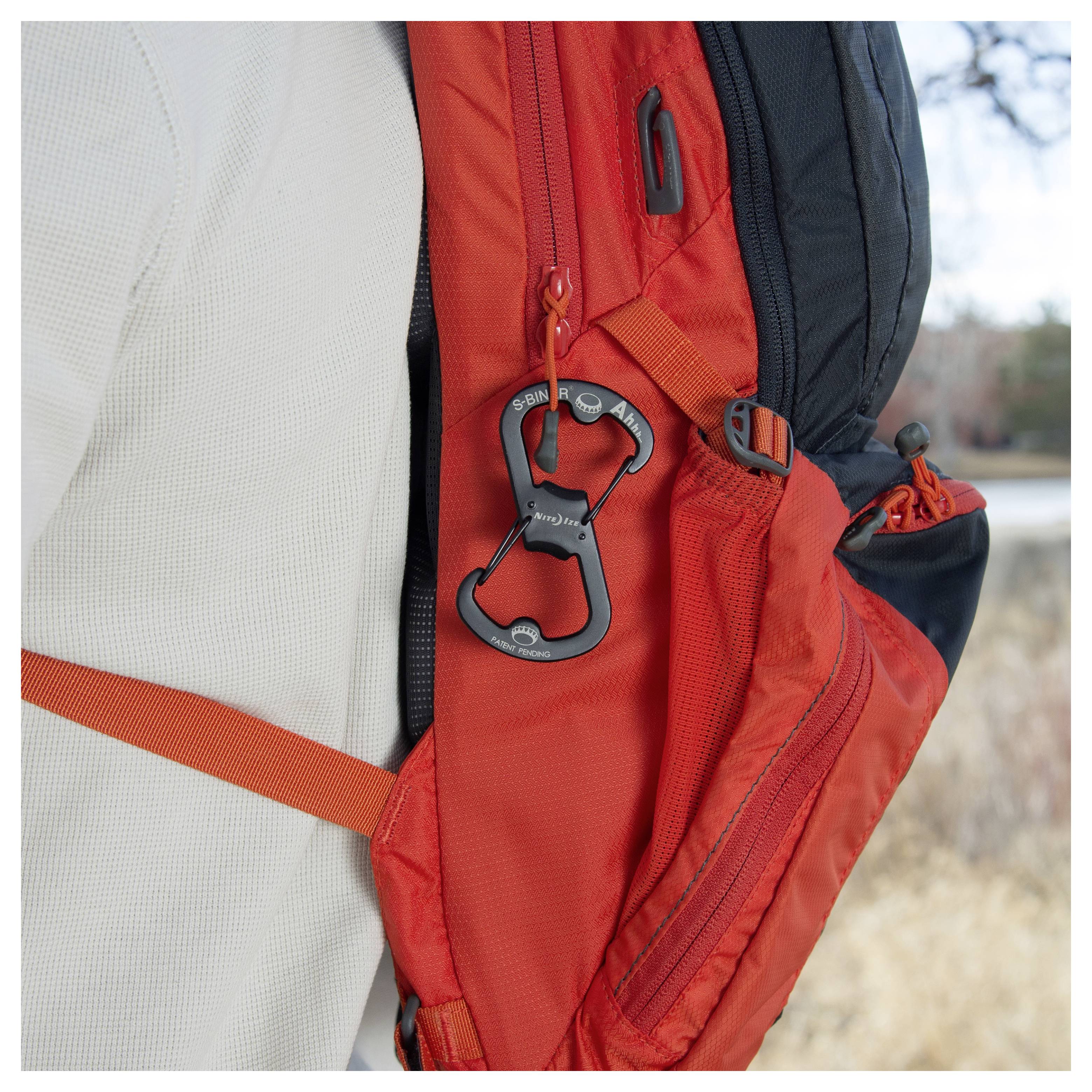 Un outil multifonction noir en forme d'ouvre-bouteille est accroché à la fermeture éclair orange d'un sac à dos porté par une personne dans un environnement extérieur.