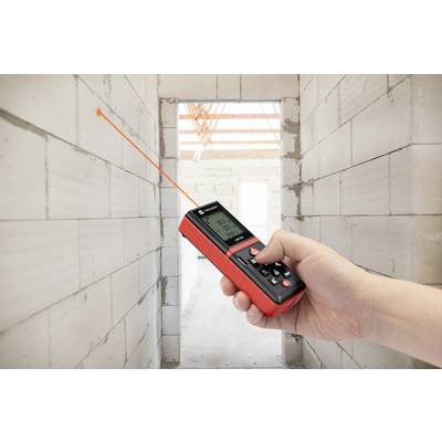 Une main tient un télémètre laser rouge et noir dans une pièce aux murs de briques blanches. Un rayon laser pointe vers le mur éloigné.