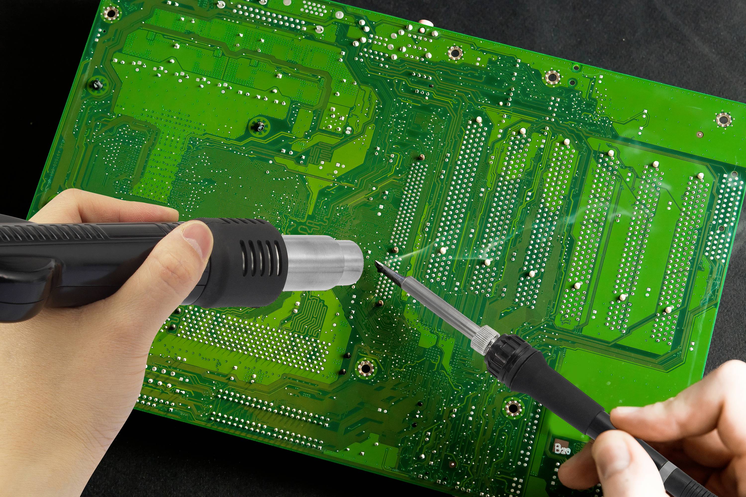 Deux mains tiennent un fer à souder et un pistolet à air chaud, tandis qu'elles travaillent sur un circuit imprimé vert.
