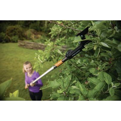 Une personne portant un haut de couleur lilas utilise un grand sécateur à longue portée pour couper des branches élevées d'un arbre vert dans un jardin.
