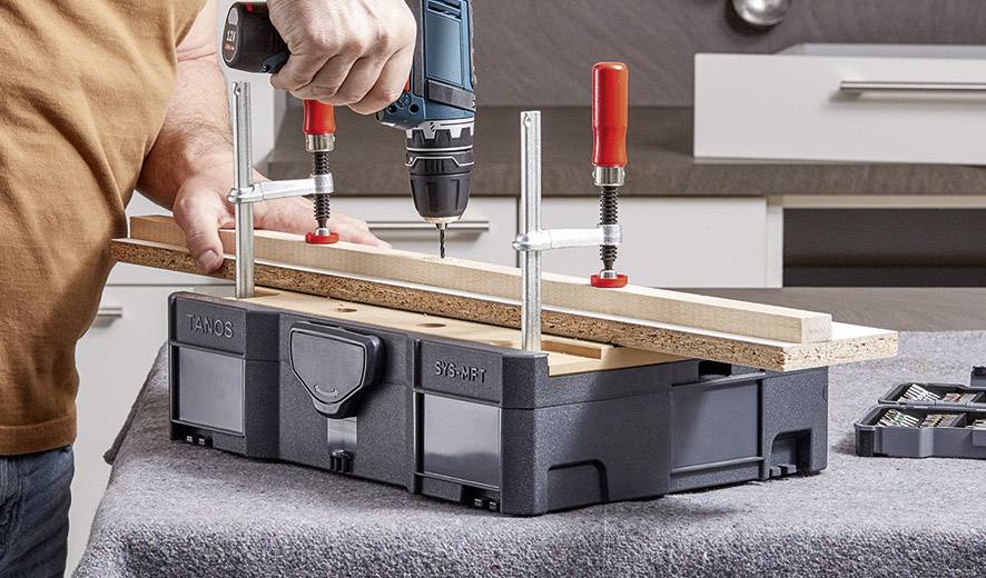 Une personne fore un morceau de bois avec une perceuse sans fil bleue, maintenu par des pinces sur un établi.