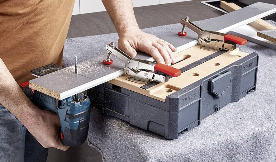 Une personne scie une planche en bois avec une scie électrique, fixée sur un établi à l'aide de pinces. Des copeaux de bois sont visibles.