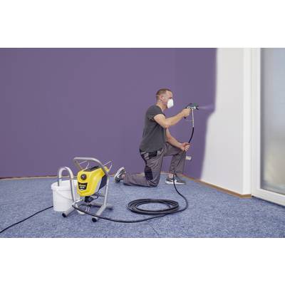 Un homme est agenouillé sur un sol en moquette et peint un mur en violet à l'aide d'un pistolet de pulvérisation. Il porte un masque de protection respiratoire.