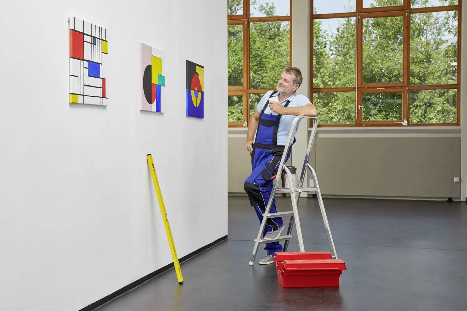 Un homme en tenue de travail est debout sur une échelle et examine des tableaux sur un mur clair de galerie. Une boîte à outils rouge est posée au sol.