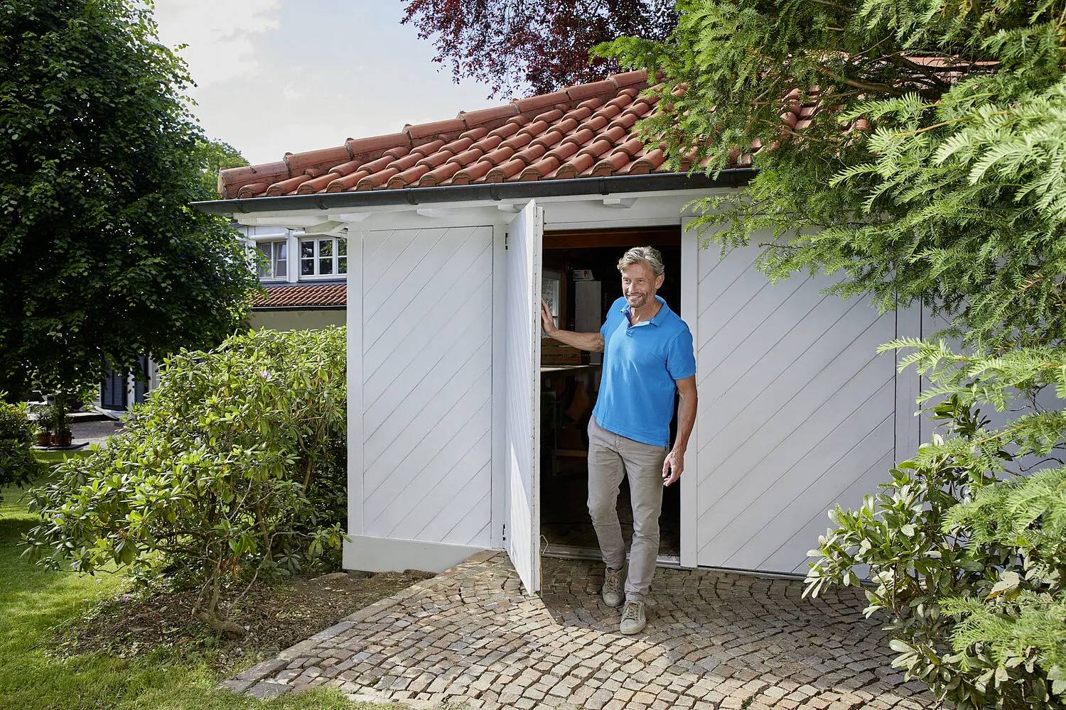 Un homme quitte une maisonnette blanche avec des briques rouges, sur un chemin pavé, entouré de buissons et d'arbres verts.