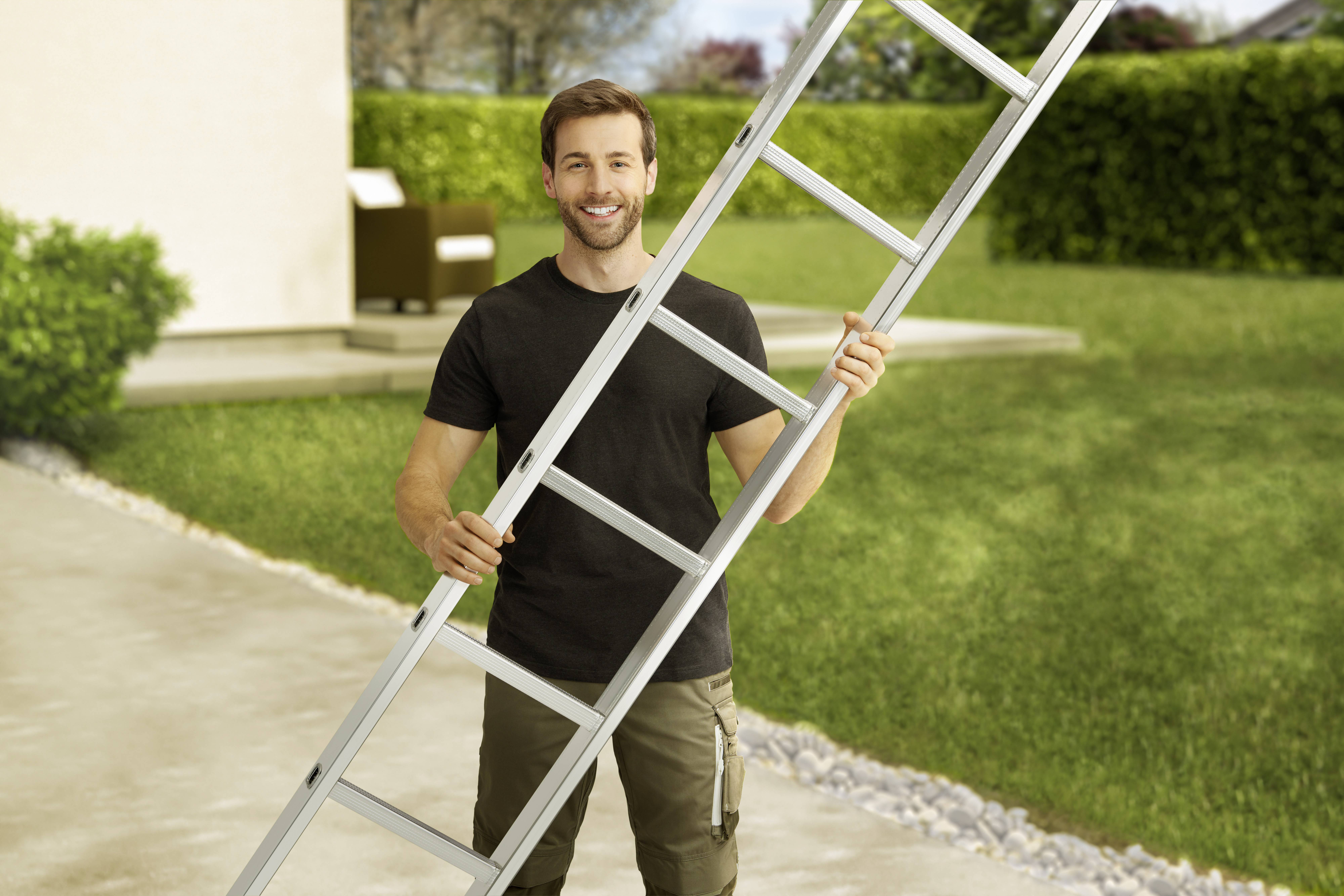 Un homme se tient à l'extérieur et tient une échelle. Un jardin et un bâtiment sont visibles en arrière-plan.