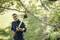 Un homme coupe des branches avec un sécateur télescopique dans un jardin. Il porte une chemise bleu foncé et se concentre sur son travail.