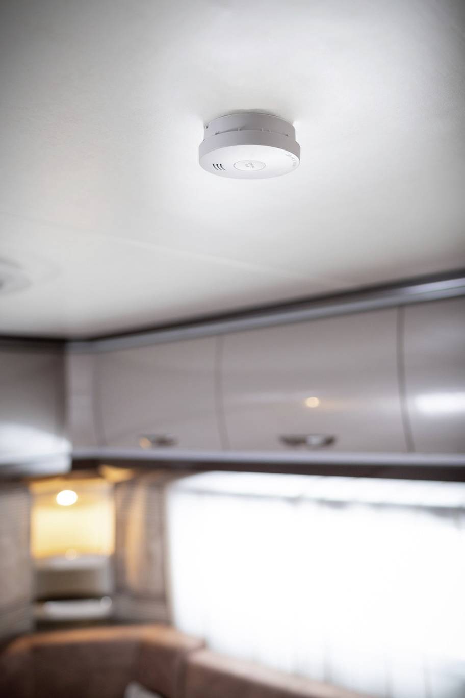 Détecteur de fumée au plafond d'un camping-car, entouré d'armoires et d'une fenêtre éclairée en arrière-plan.