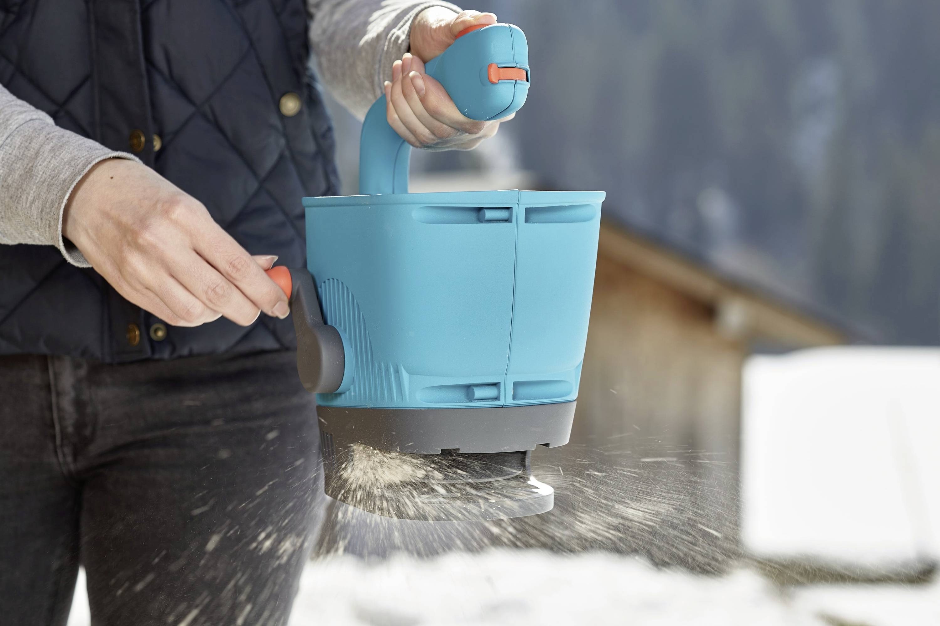 Une personne tient un dispositif bleu d'épandage, répand du sable ou du sel sur une surface enneigée. L'appareil est utilisé à l'extérieur.
