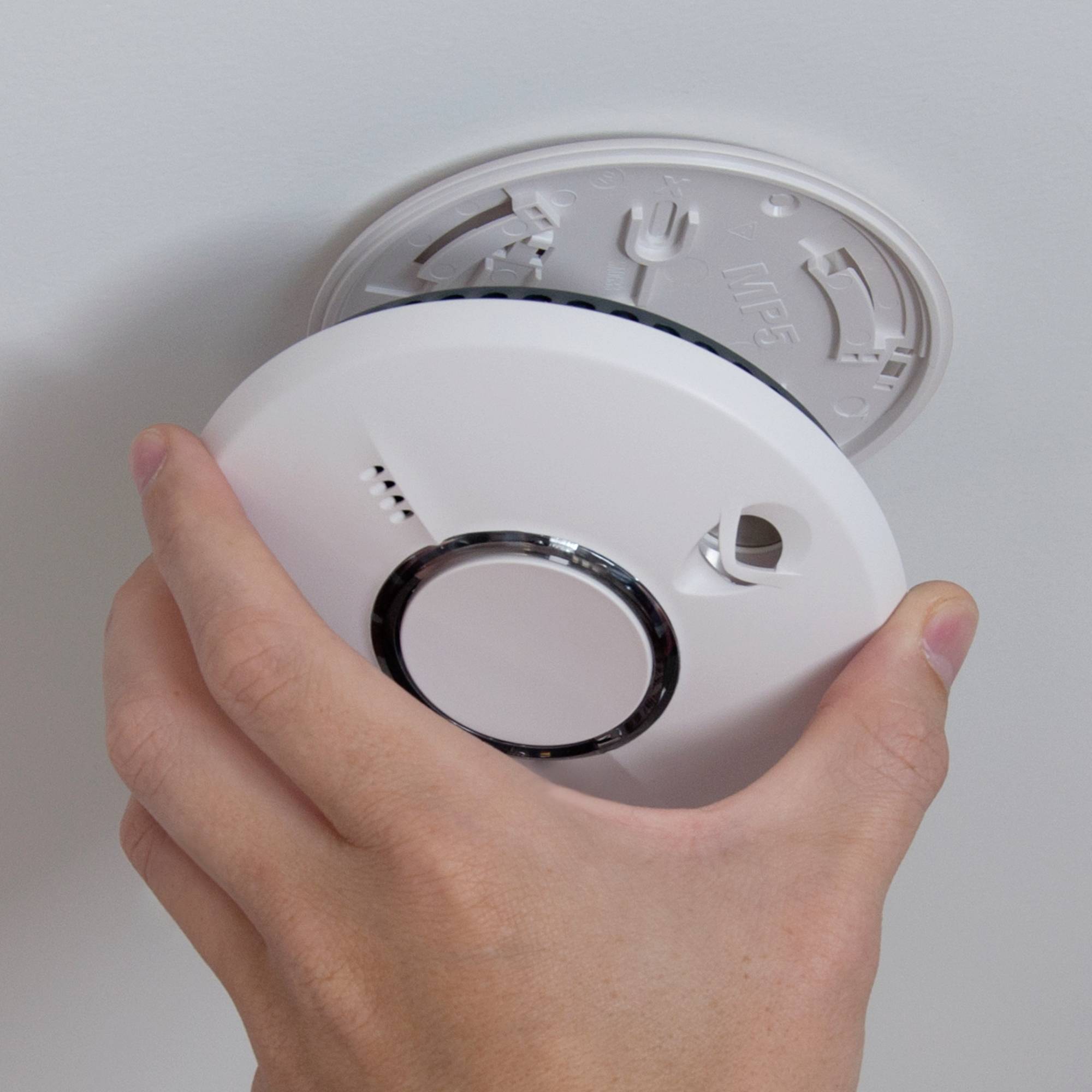 Une personne installe un détecteur de fumée blanc sur un support de plafond, veillant à la sécurité de la maison. L'appareil présente un design circulaire et dispose d'un bouton de test.