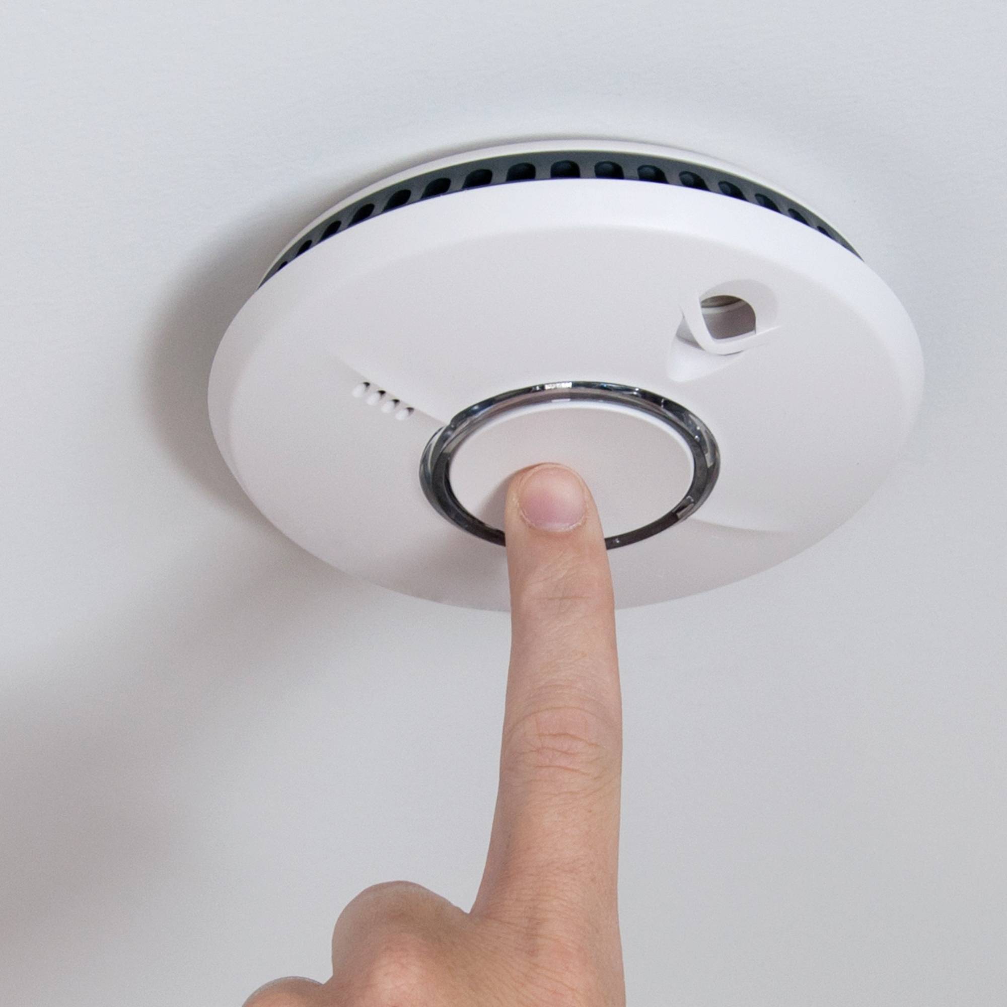 Une main appuyant sur le bouton de test d'un détecteur de fumée monté au plafond.