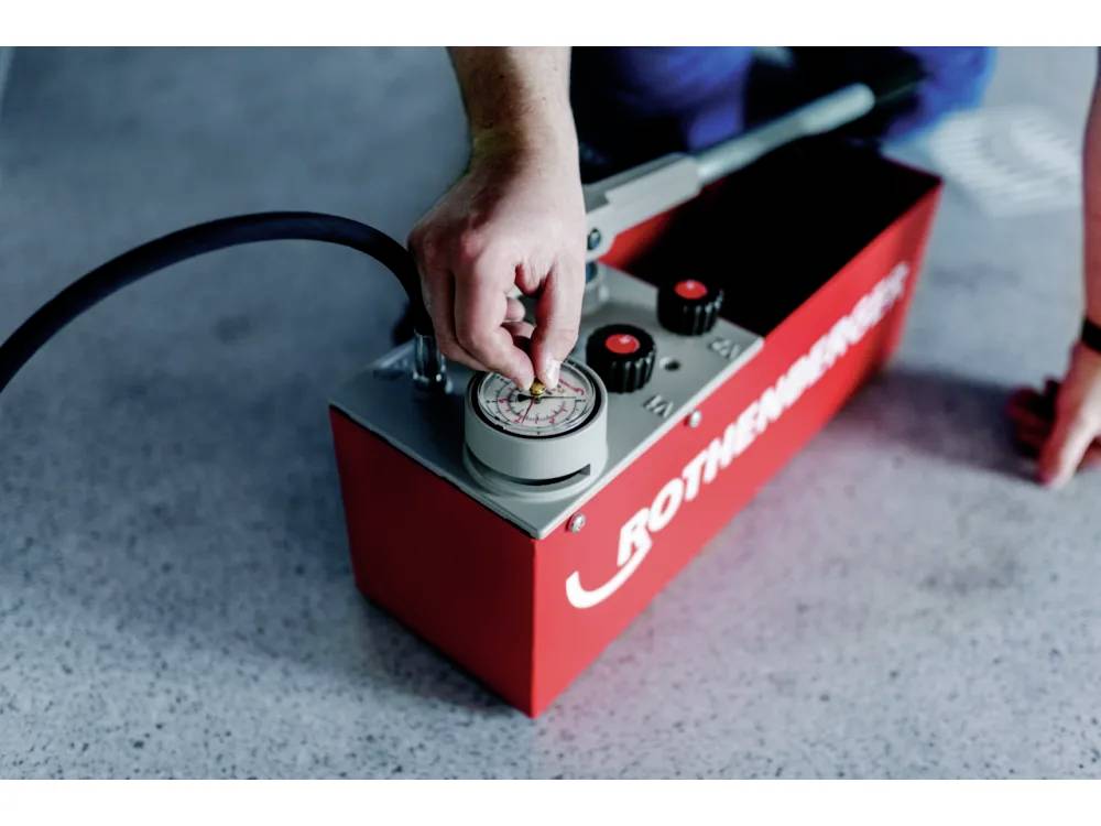 Une personne ajustant un manomètre sur un outil hydraulique rouge, posé sur une surface grise mouchetée. L'outil est doté d'un tuyau qui y est connecté.