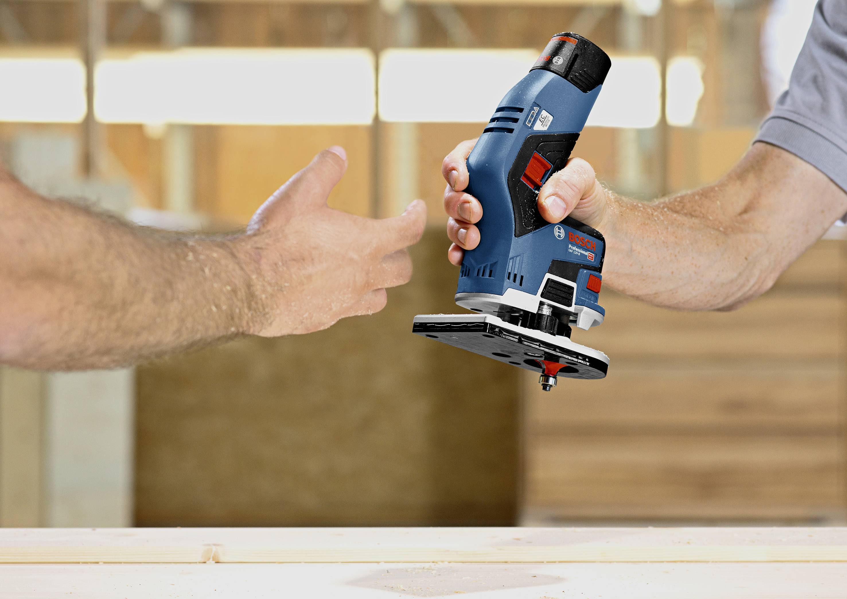 Une personne tend un outil électrique bleu à une autre personne dans un atelier. Des structures en bois sont visibles en arrière-plan.