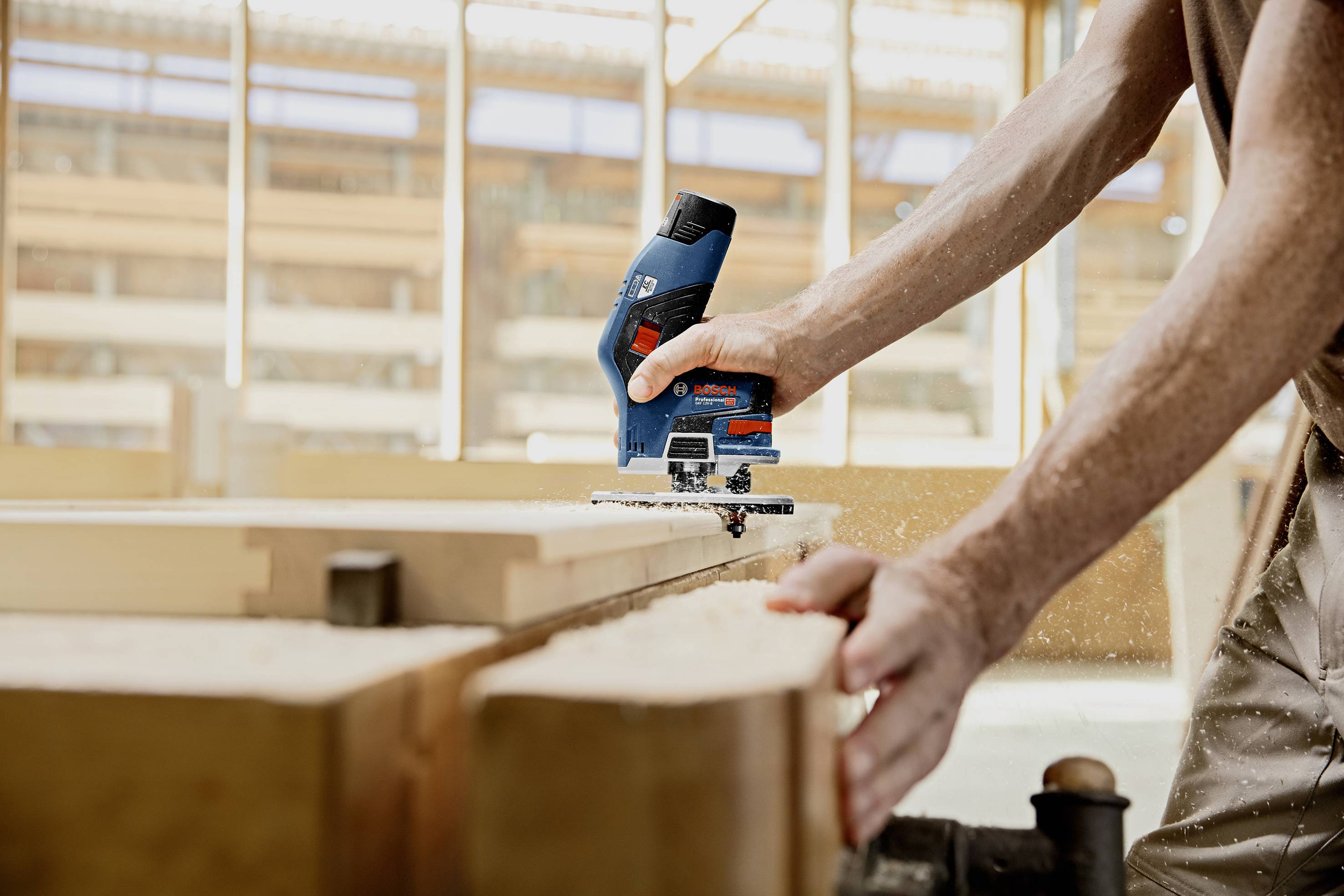 Une personne scie précisément du bois avec une scie sauteuse bleue dans un atelier lumineux. Des copeaux de bois volent dans les airs.
