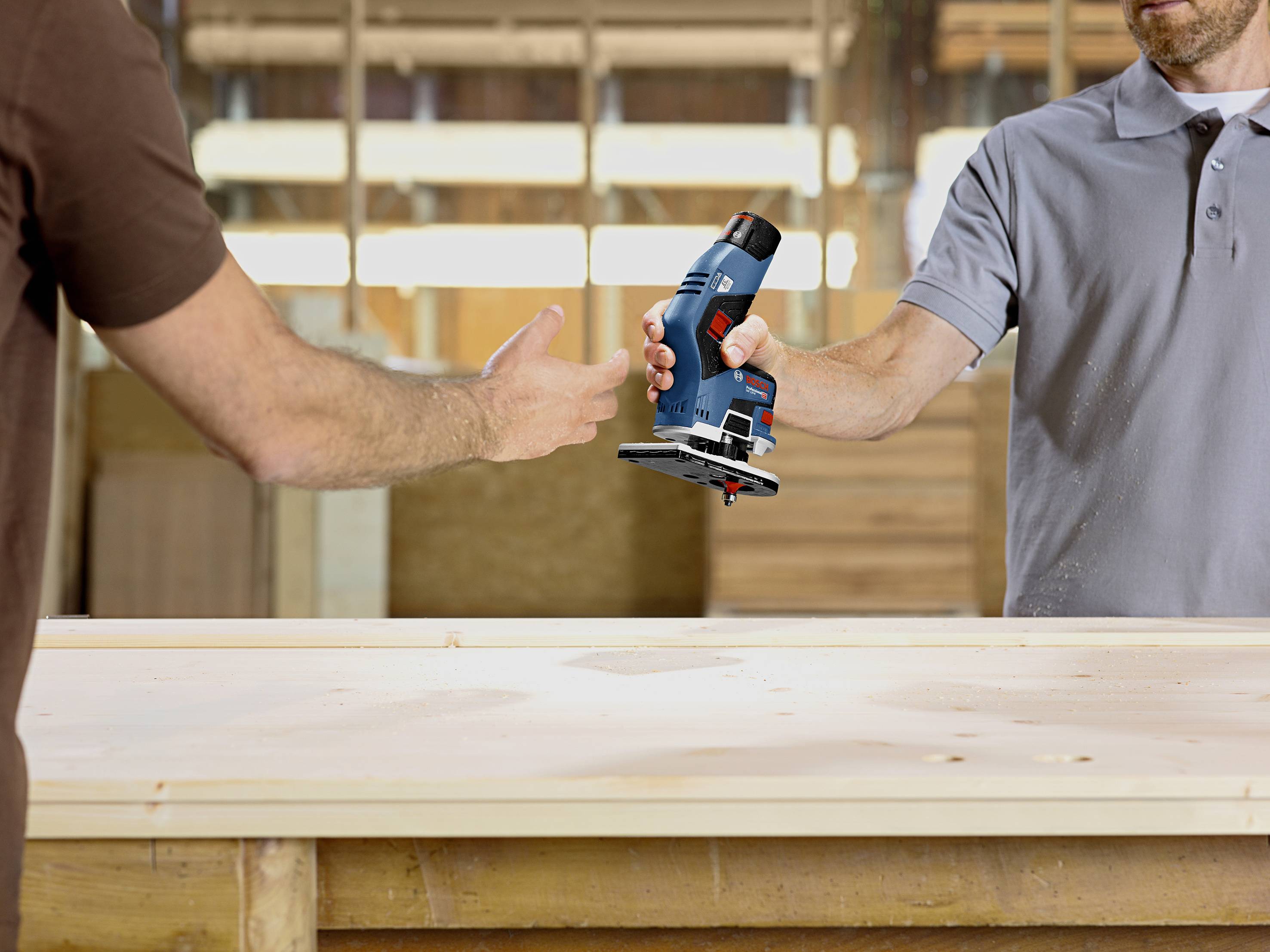 Deux personnes se passent une fraiseuse électrique par-dessus un établi dans un environnement d'atelier.