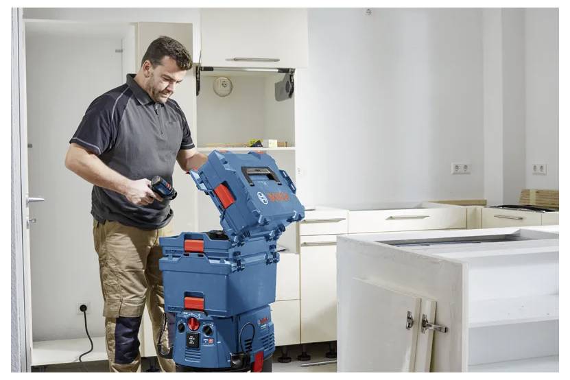 Une personne dans un environnement domestique utilise un aspirateur bleu Bosch avec plusieurs compartiments de rangement, entouré d'armoires blanches.