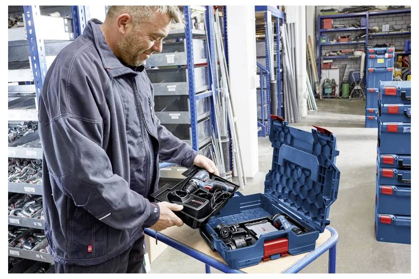 Un homme dans un atelier ouvre un coffret à outils bleu et rouge sur une table, en étalant son contenu. Des étagères remplies d'outils et de boîtes l'entourent.