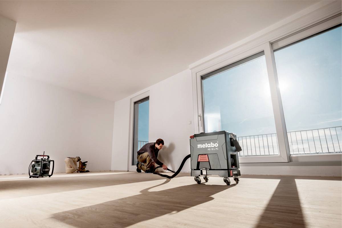 Une personne pose un plancher en bois dans une pièce lumineuse avec de grandes fenêtres. Un aspirateur de 'Metabo' est posé à côté de la personne.