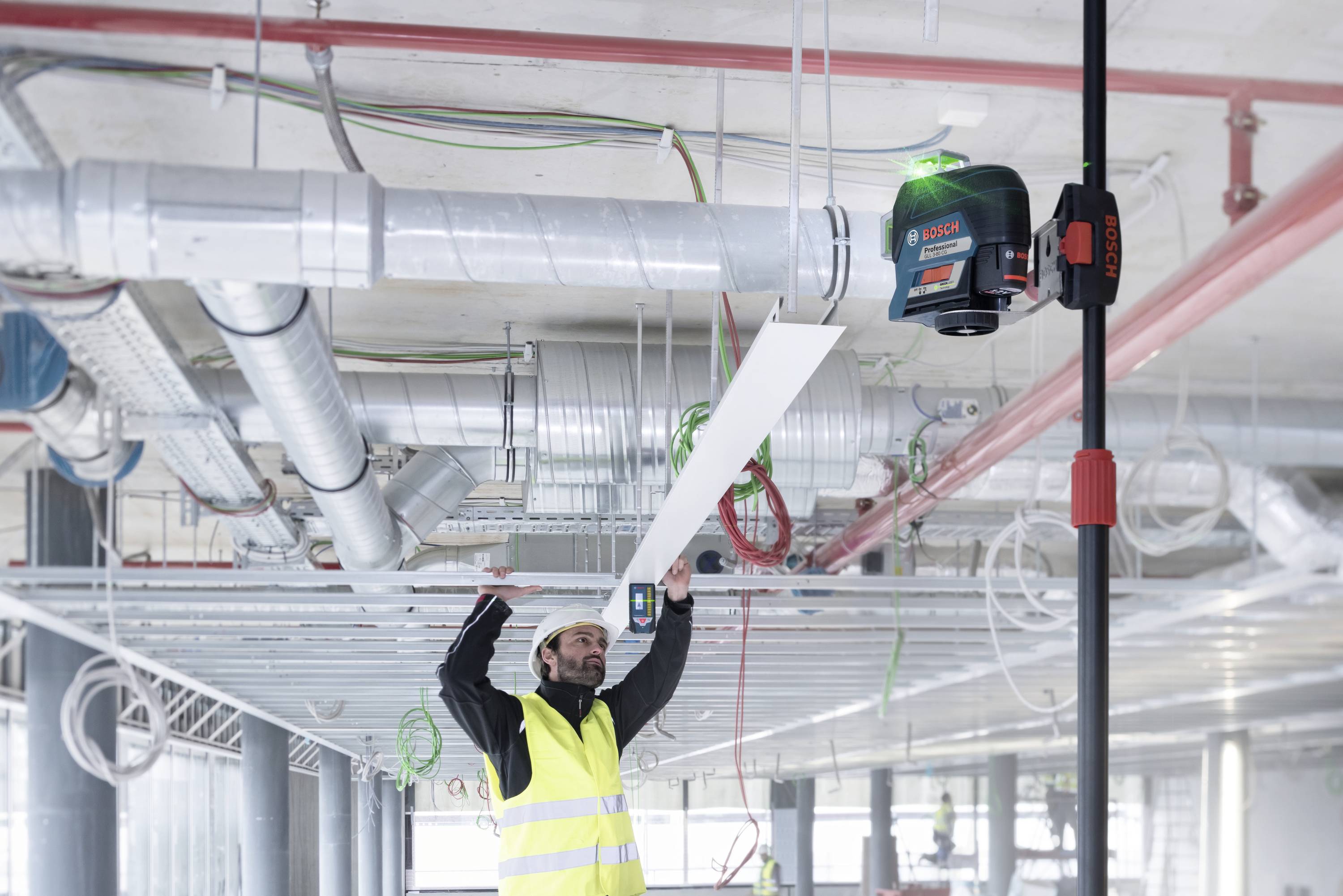 Un ouvrier aligne une plaque métallique à l'aide d'un niveau laser vert, entouré de tuyaux et de câbles sur un chantier de construction.