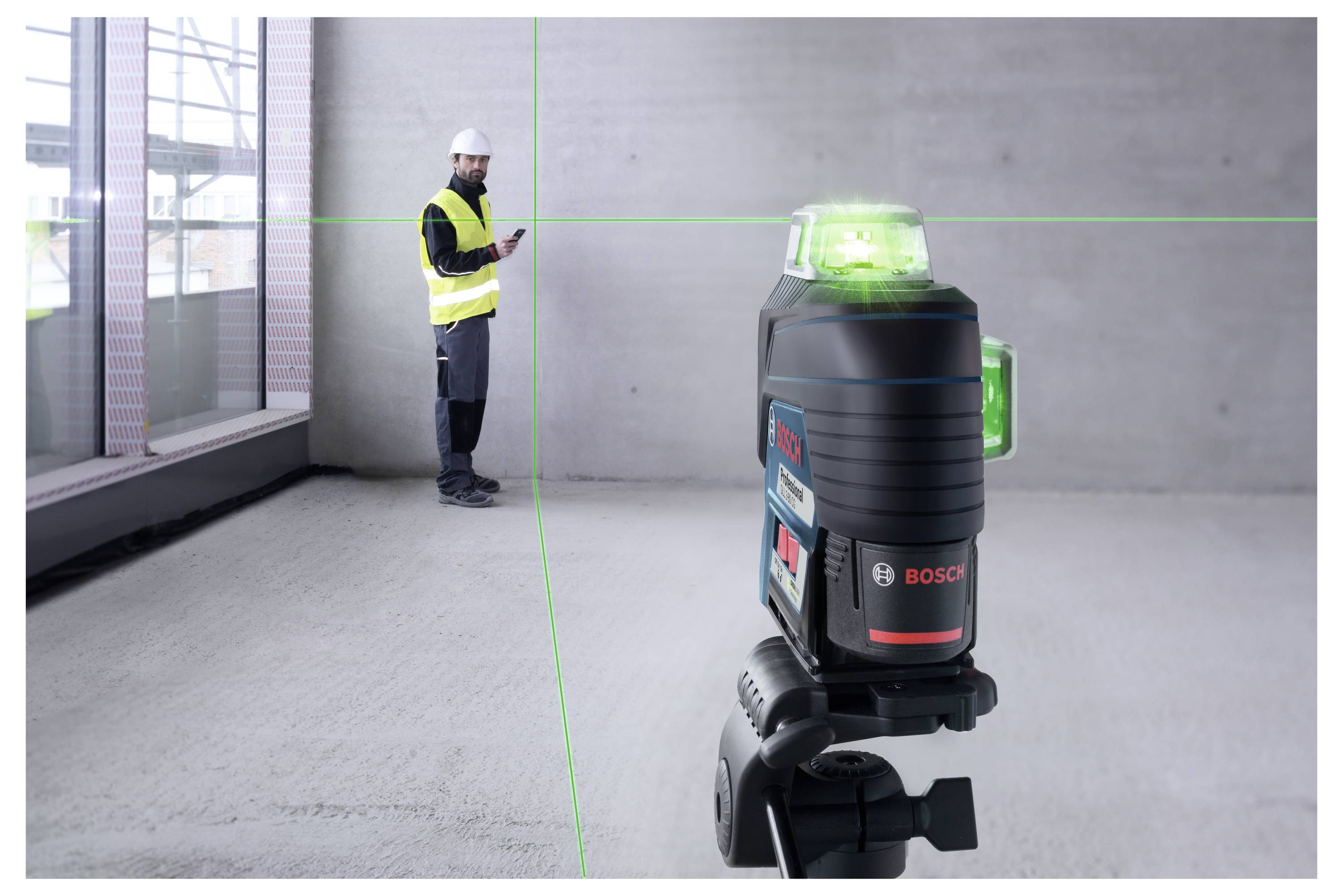 Un ouvrier en tenue de protection utilise un smartphone pendant qu'un appareil de nivellement laser projette une ligne verte sur un mur en béton.