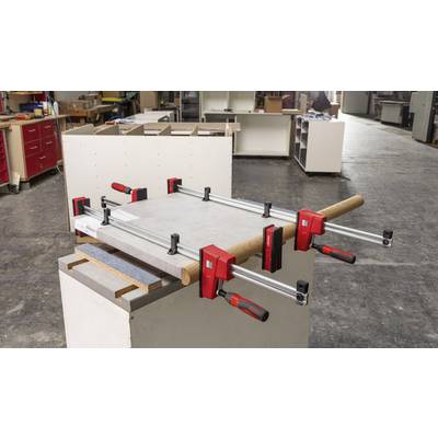 Un établi avec plusieurs pinces fixant un morceau de bois. À l'arrière-plan, d'autres outils et meubles sont visibles dans un atelier.
