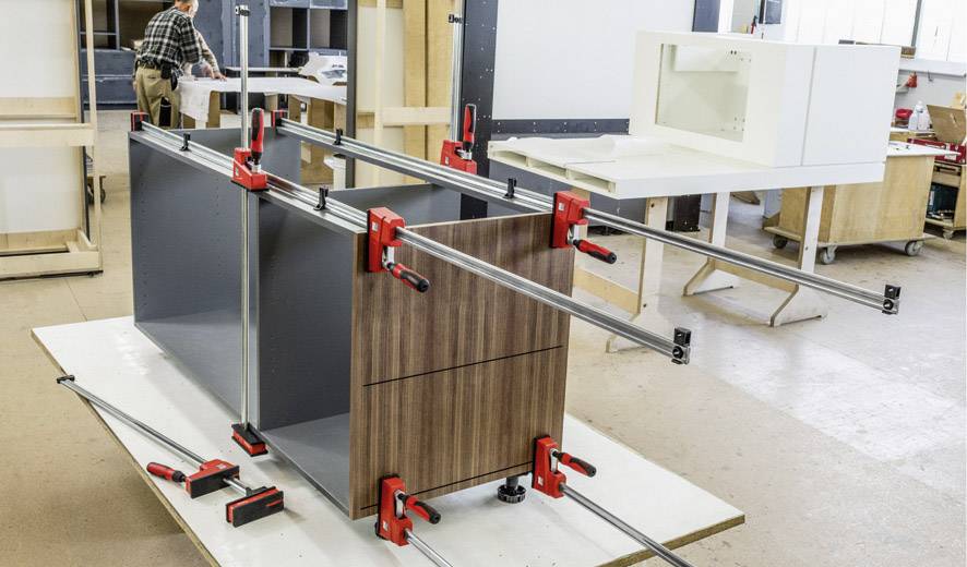 Un atelier de menuiserie avec un ouvrier en arrière-plan. Une pièce de mobilier au centre est maintenue par des serre-joints pour séchage.