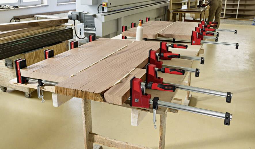 Des planches de bois sont fixées avec des serre-joints rouges sur une table de travail dans un atelier. Des machines sont visibles en arrière-plan.