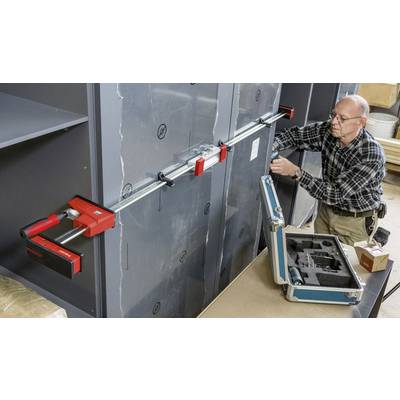 Un homme fixe un coffre-fort avec une grande pince rouge. À côté de lui, une mallette à outils ouverte contient du matériel supplémentaire.