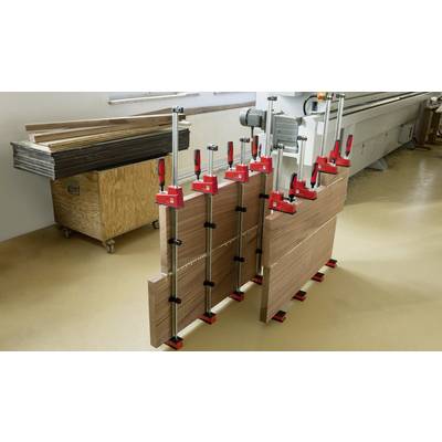 Des planches de bois sont pressées ensemble à l'aide de grandes serre-joints dans un atelier. Une machine est visible en arrière-plan.