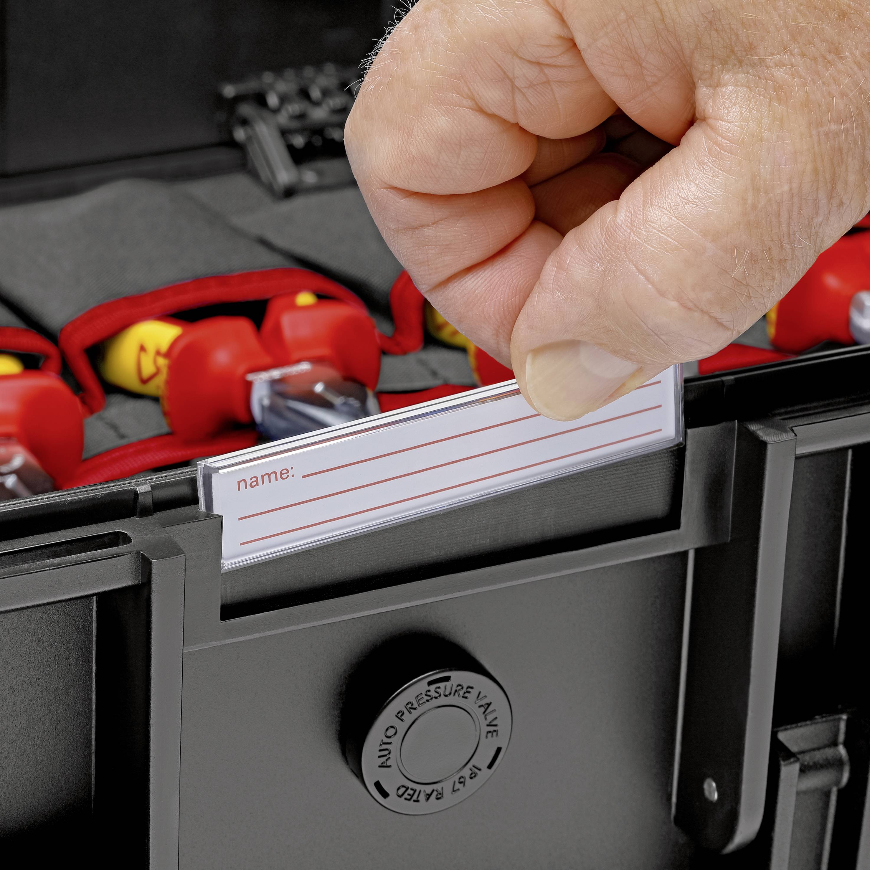 Une main insère une carte descriptible dans un compartiment nominatif d'une valise noire. En arrière-plan, des éléments rouges avec des clips jaunes sont visibles.