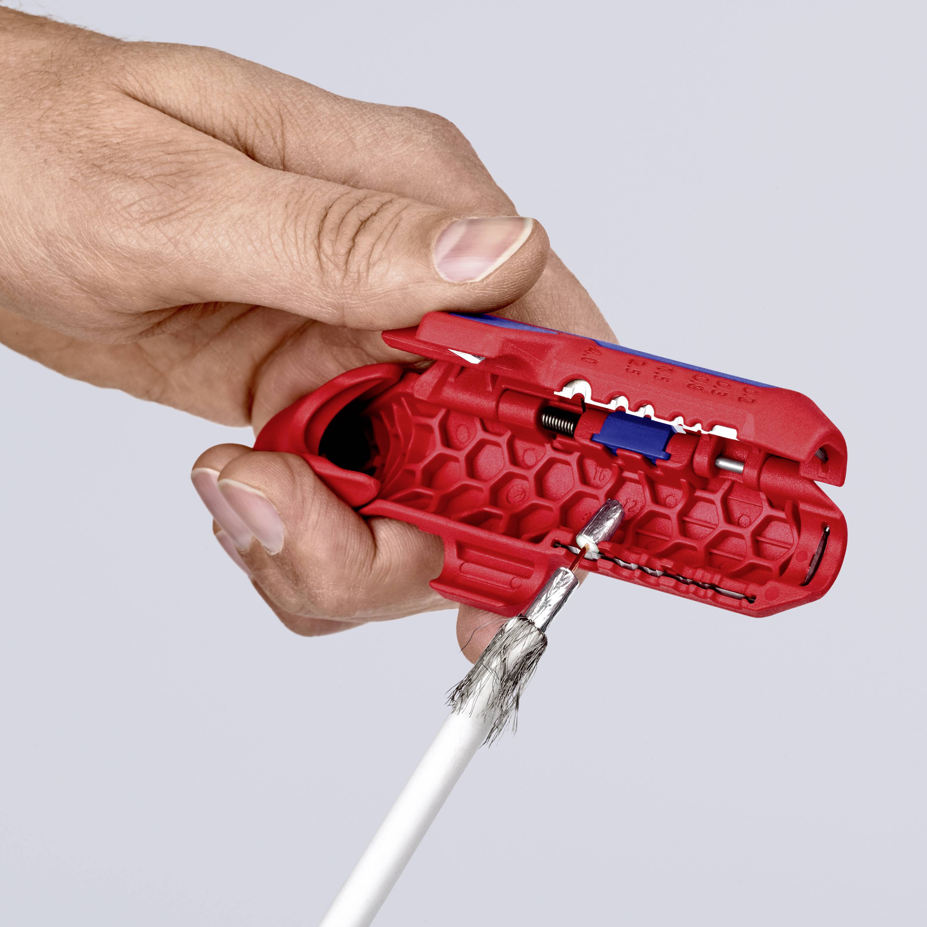 Une main tient un outil rouge utilisé pour dénuder un câble blanc.