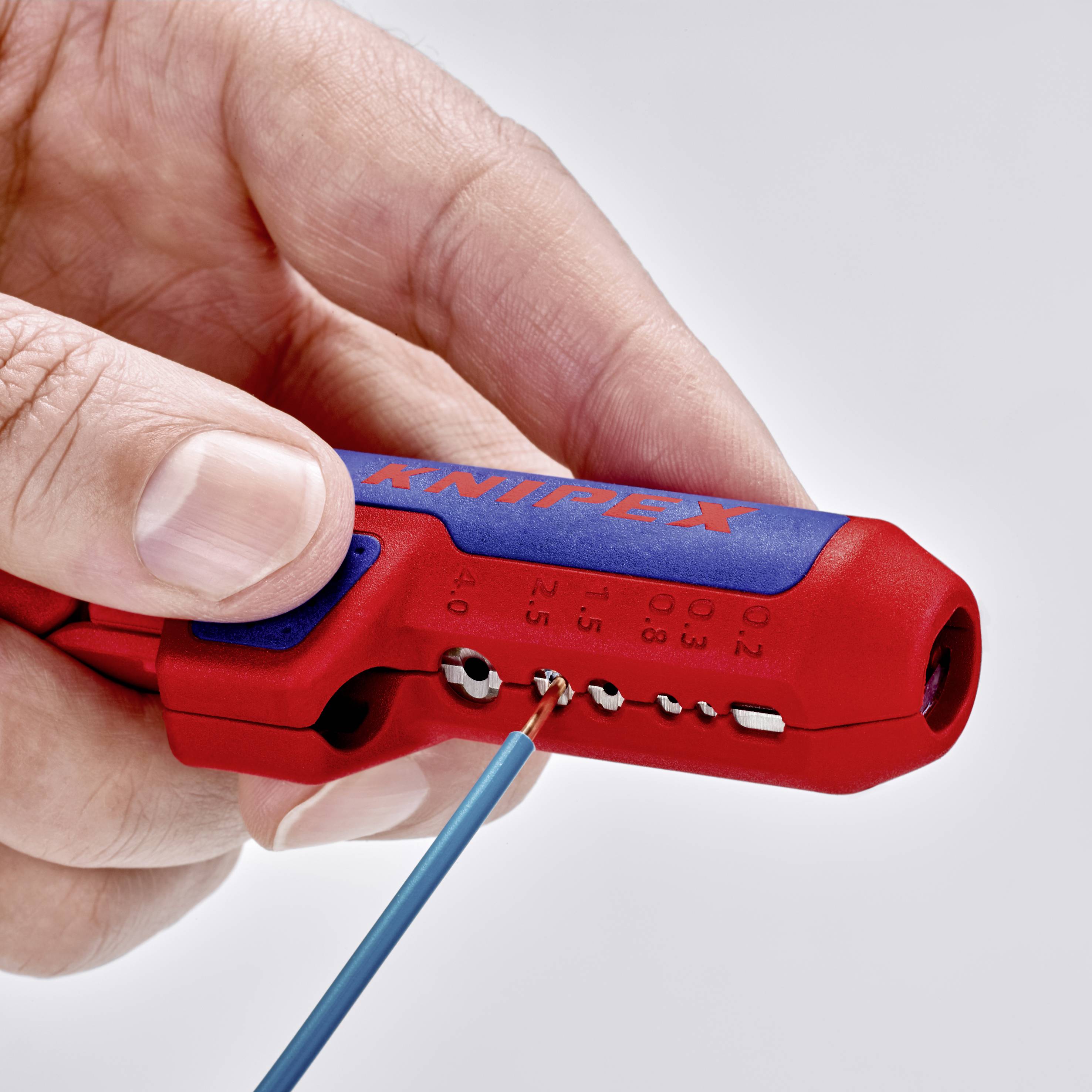 Une main tient un outil de dénudage rouge et bleu de la marque Knipex, utilisé pour dénuder un câble bleu.