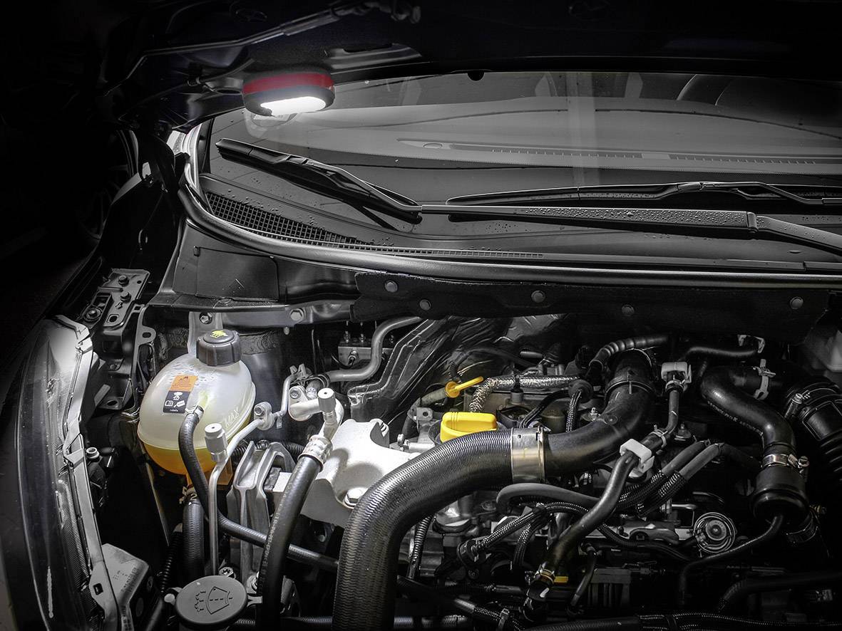 Un compartiment moteur ouvert d'une voiture avec des tuyaux, des câbles et un vase d'expansion visible à gauche. L'espace est éclairé depuis le haut.