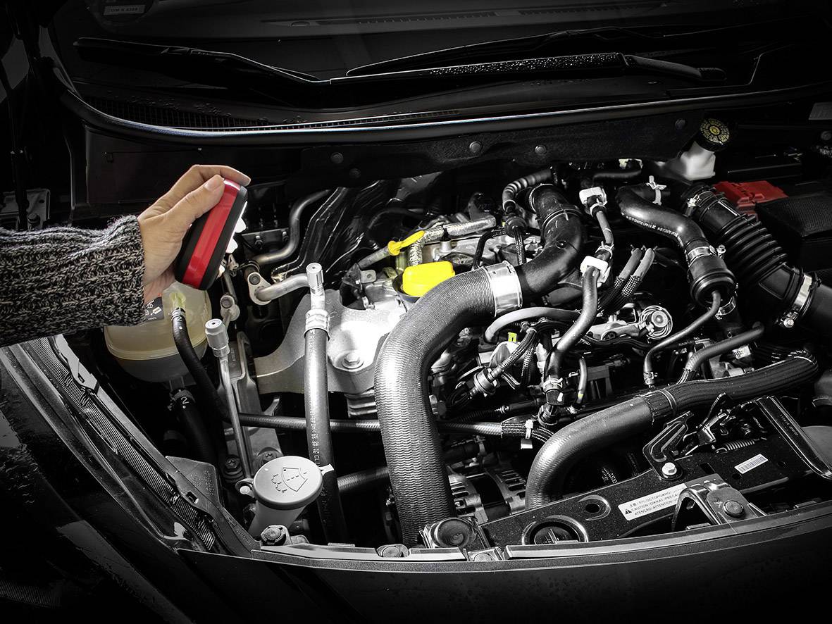 Une main tient un manuel de moteur ouvert au-dessus d'un moteur de voiture complexe avec des tuyaux, des câbles et des réservoirs de liquide visibles.