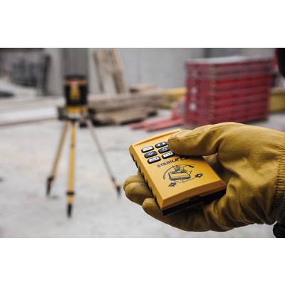 Une main gantée tient un dispositif de commande jaune, avec un trépied flou visible en arrière-plan.