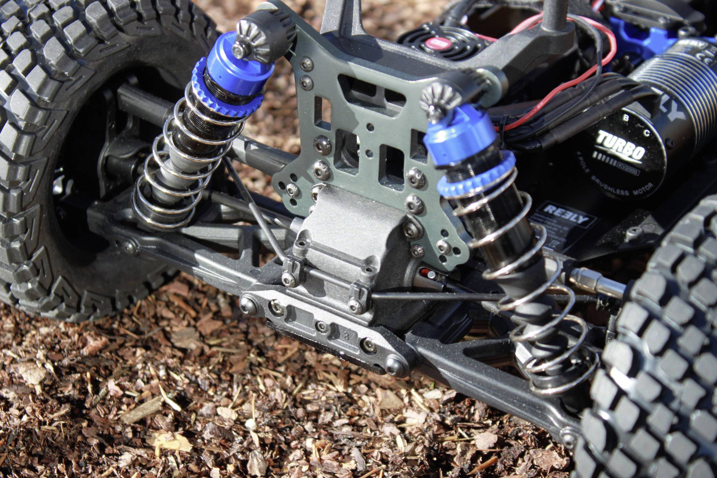 Suspension d'une voiture télécommandée avec des amortisseurs bleus et des pneus à gros crampons, positionnée sur un sol de copeaux de bois.